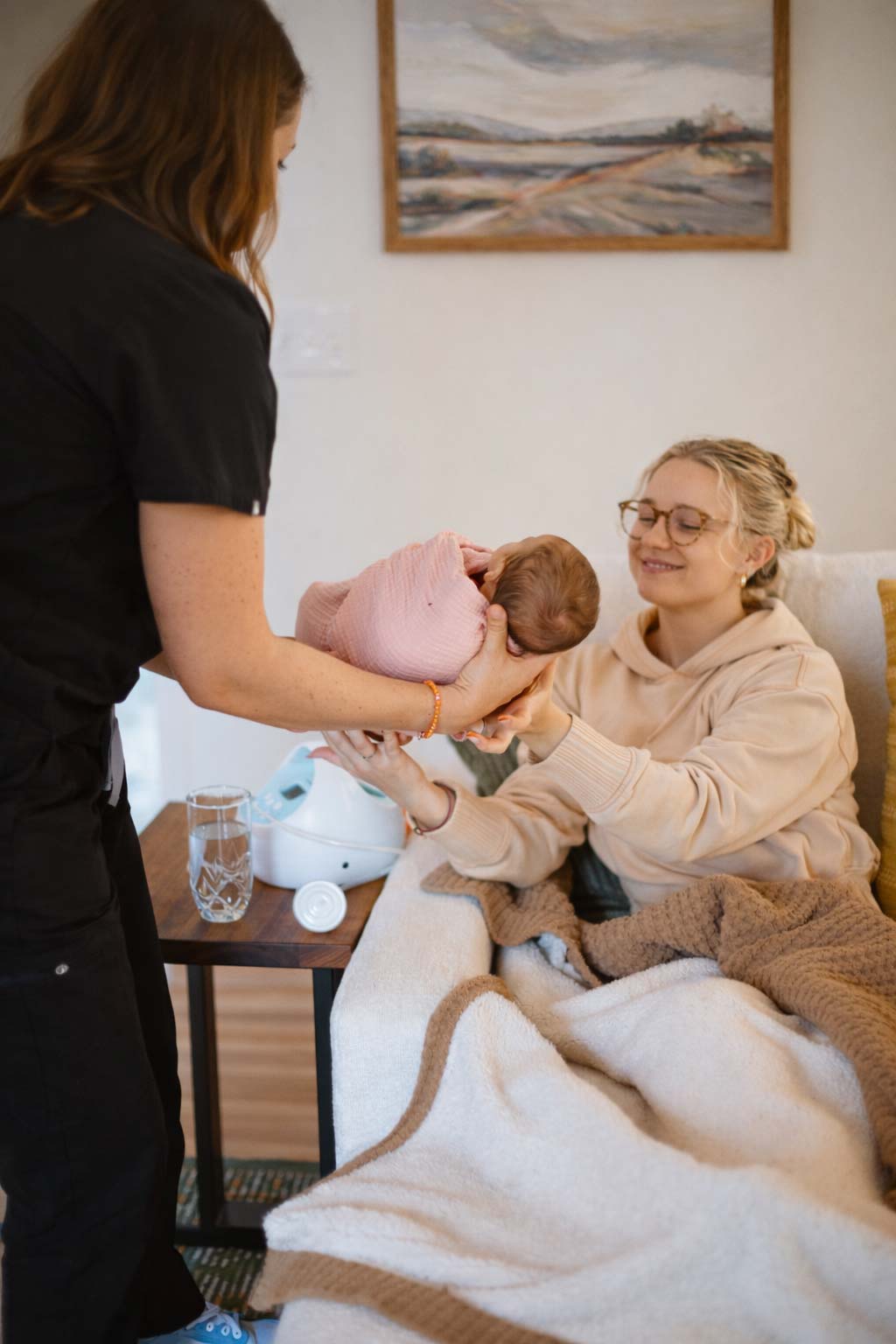 A nurse handing a swaddled baby to a mother resting on a sofa under blankets