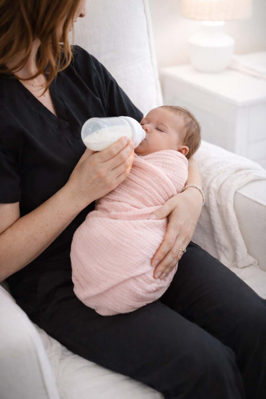 A nurse in black scrubs bottle feeding a swaddled baby in an armchair
